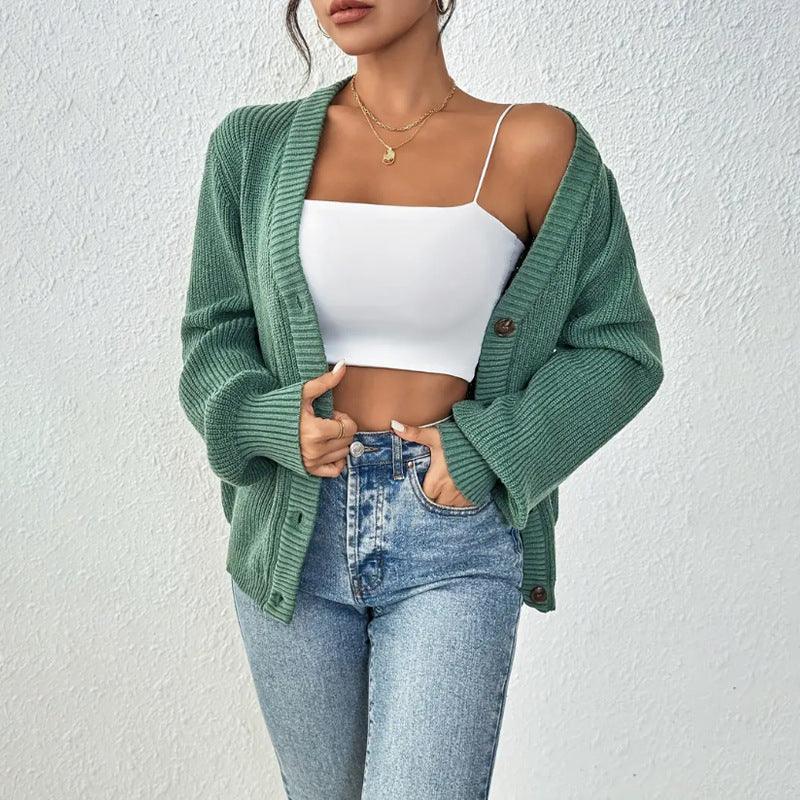 Woman wearing green knit cardigan, white crop top, and high-waisted blue jeans against white wall