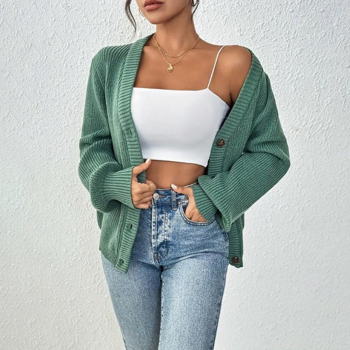 Woman wearing green knit cardigan, white crop top, and high-waisted blue jeans against white wall