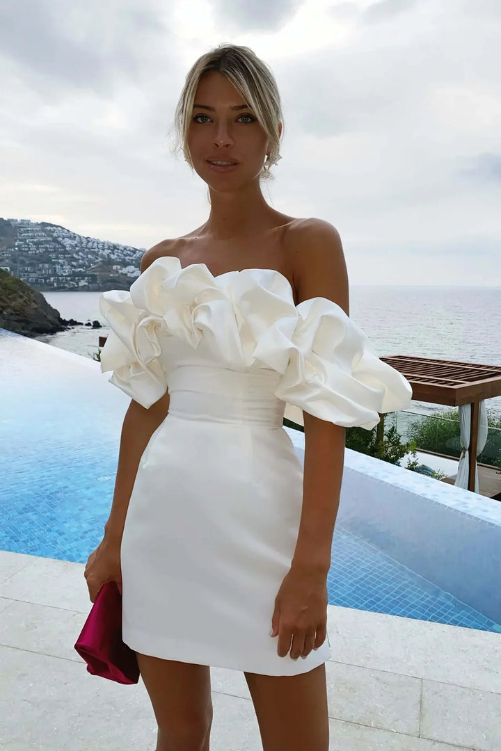 Woman wearing a white off-shoulder ruffled mini dress standing by infinity pool with ocean view