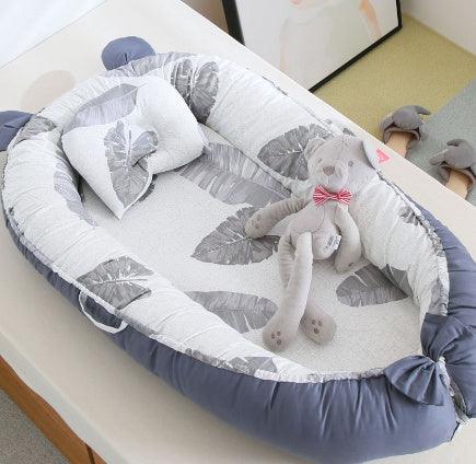 Baby nest bed with gray and white leaf pattern, small pillow, and beige teddy bear with striped bow