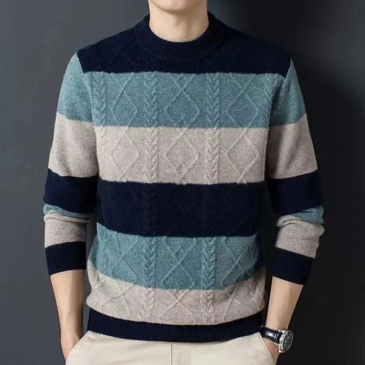Man wearing a striped cable-knit sweater in blue and beige tones with beige pants against dark background