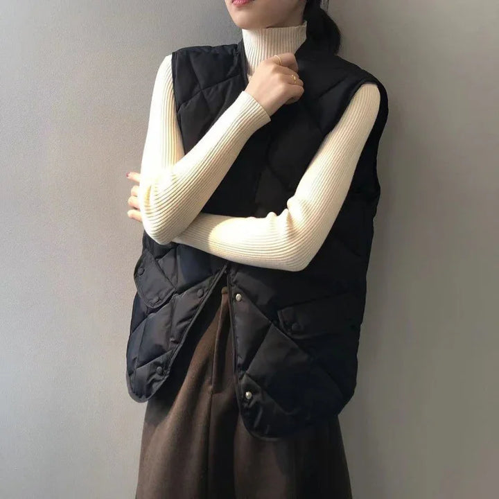 woman wearing black quilted sleeveless vest over cream ribbed turtleneck sweater and brown pants against plain wall