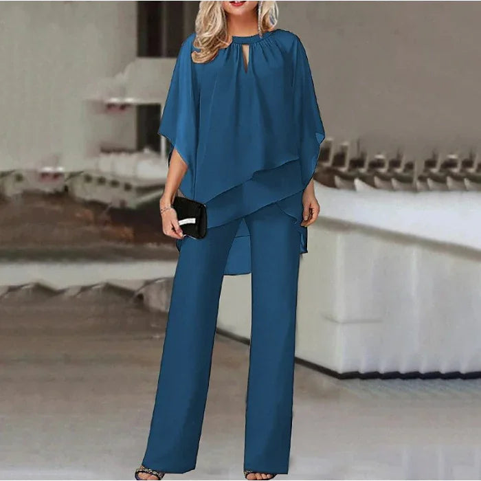 Woman in elegant blue layered tunic and wide-leg pants holding black clutch indoors