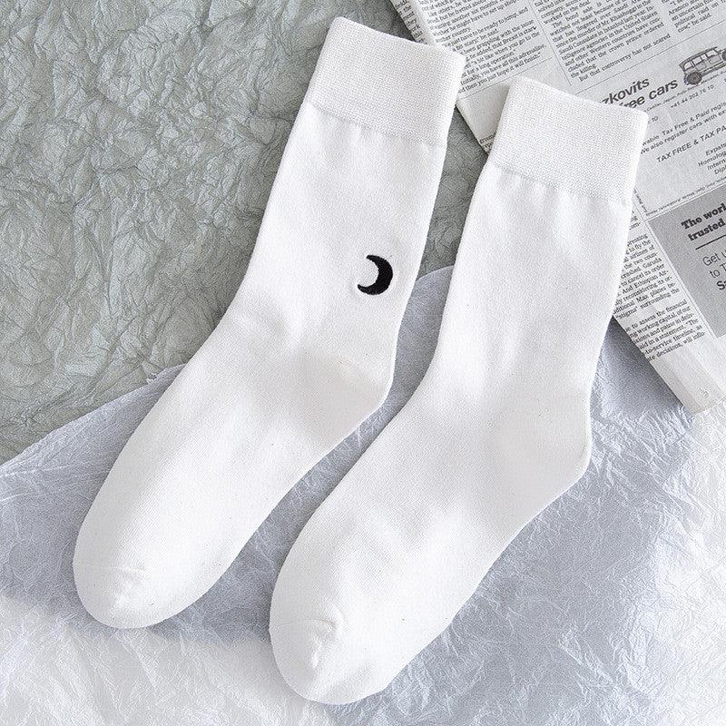 Pair of white crew socks with black crescent moon design, laid on textured gray and white surface with newspaper