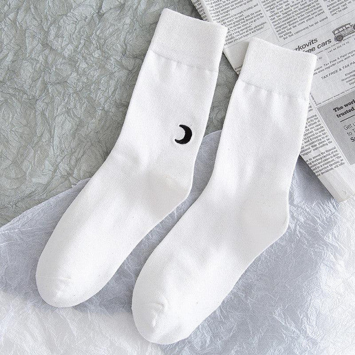Pair of white crew socks with black crescent moon design, laid on textured gray and white surface with newspaper