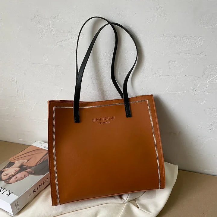 Brown leather tote bag with black handles and white stitching, branded JINGPINPIJU FASHION, on beige surface