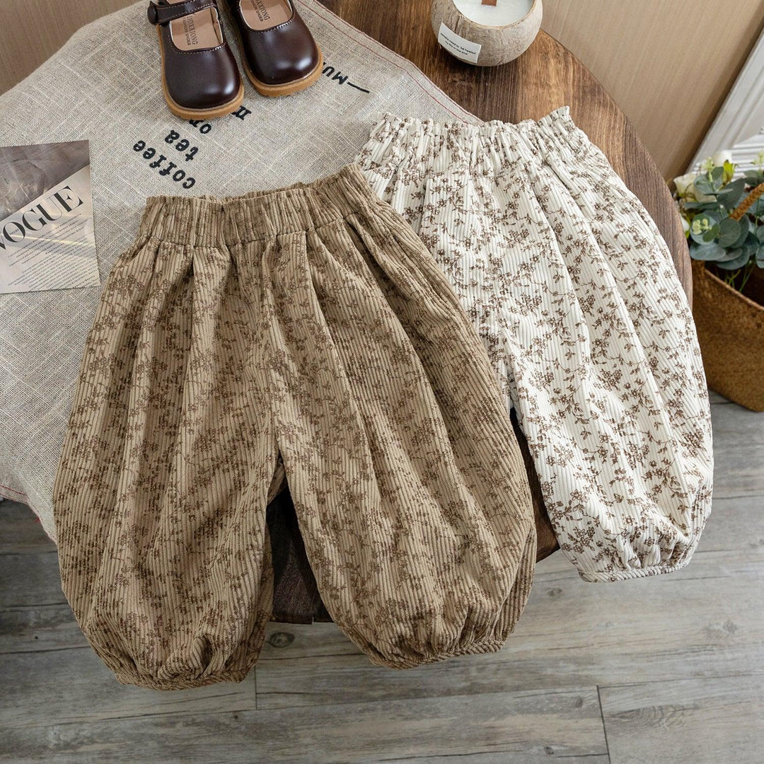 Two pairs of floral-patterned toddler balloon pants, one beige and one white, on a wooden table with brown shoes and decor