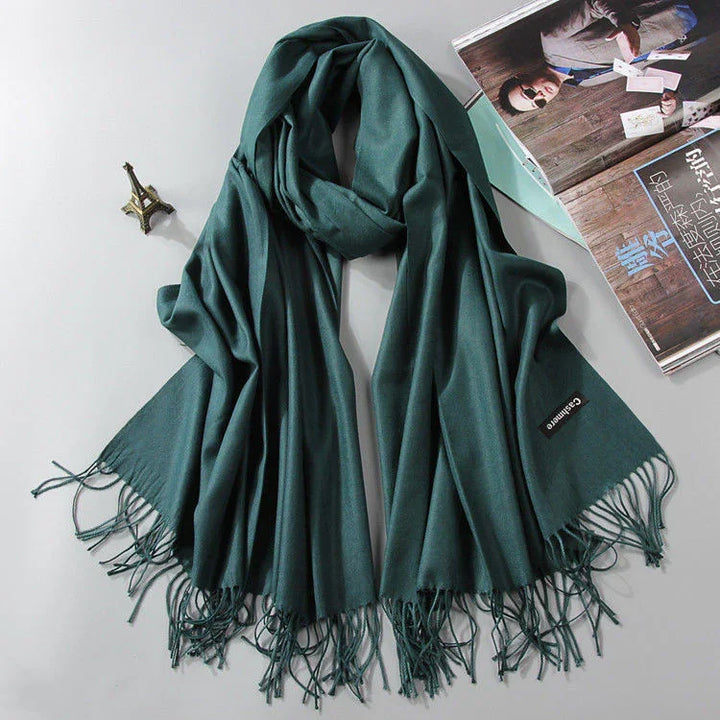 Dark green cashmere scarf with fringes displayed on white surface, alongside small Eiffel Tower replica and open magazine