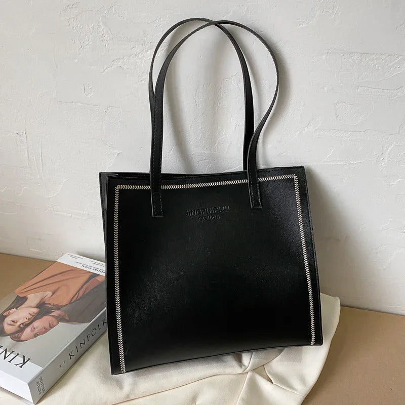 Black leather tote bag with white edge stitching and embossed logo, placed on white cloth near a book
