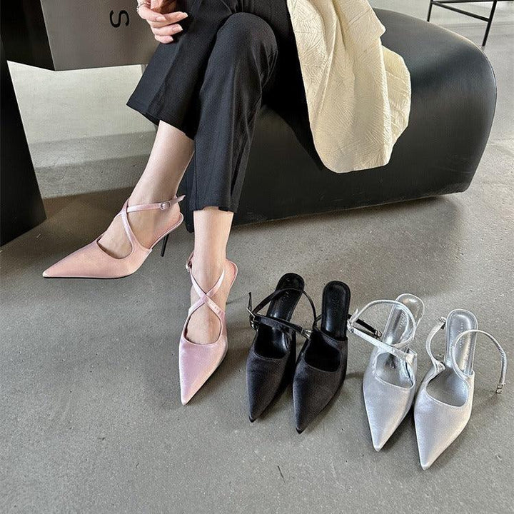 Woman wearing pink pointed-toe slingback heels with crisscross straps sitting next to black and silver heels on concrete floor