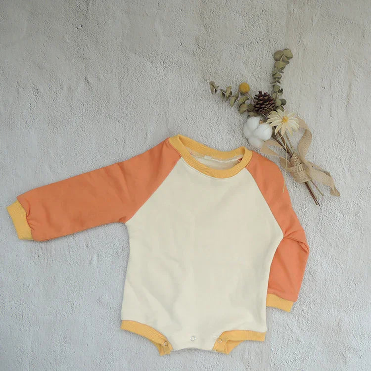 Baby onesie with orange sleeves and cream body, yellow collar and cuffs, laid on textured gray surface with dried flower arrangement