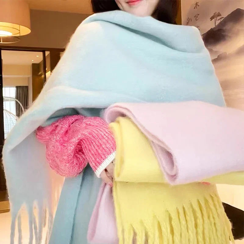 Person indoors wrapped in light blue scarf, holding yellow and pink fringed scarves, wearing pink sweater