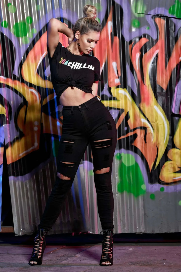 Fashionable woman in ripped black jeans, black tied crop top, and lace-up heels posing against colorful graffiti wall