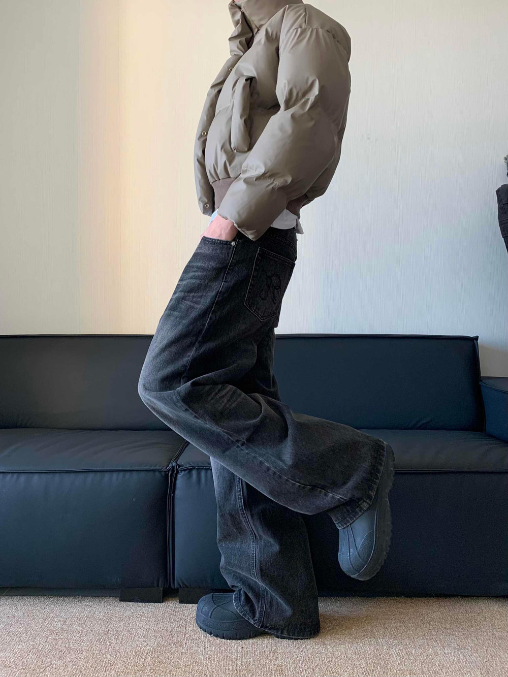 Person wearing a beige puffer jacket, black wide-leg jeans with embroidery, and black chunky shoes indoors