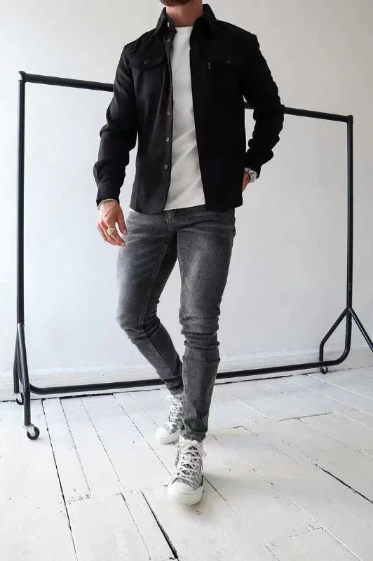 Man in black shirt, white t-shirt, gray jeans, and white sneakers posing on white wooden floor