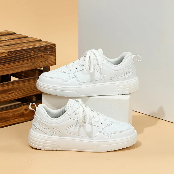 Pair of white casual sneakers with laces displayed on beige surface with wooden crate background