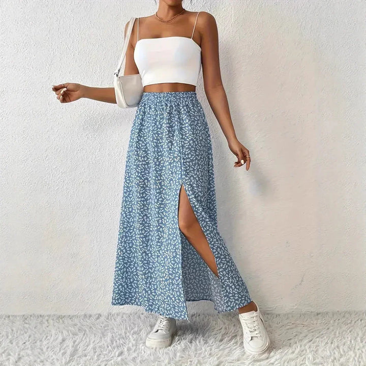 Woman wearing white crop top, blue floral slit skirt, white sneakers, posing indoors on white textured background