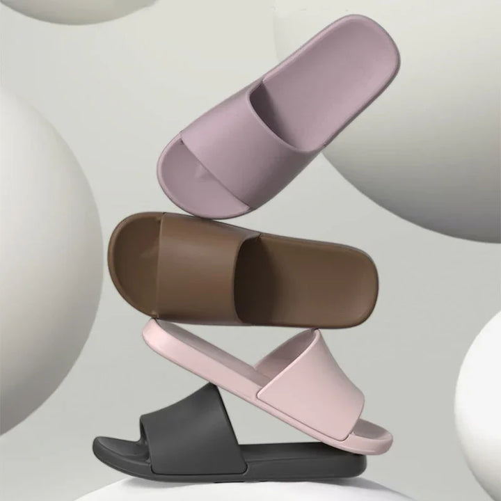 Stack of four modern slide sandals in purple, brown, pink, and black against a neutral background with large white spheres