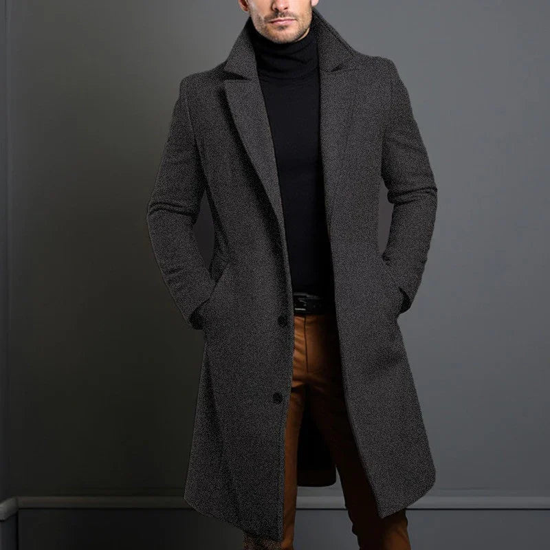 Man wearing dark gray wool coat over black turtleneck and brown pants against gray background