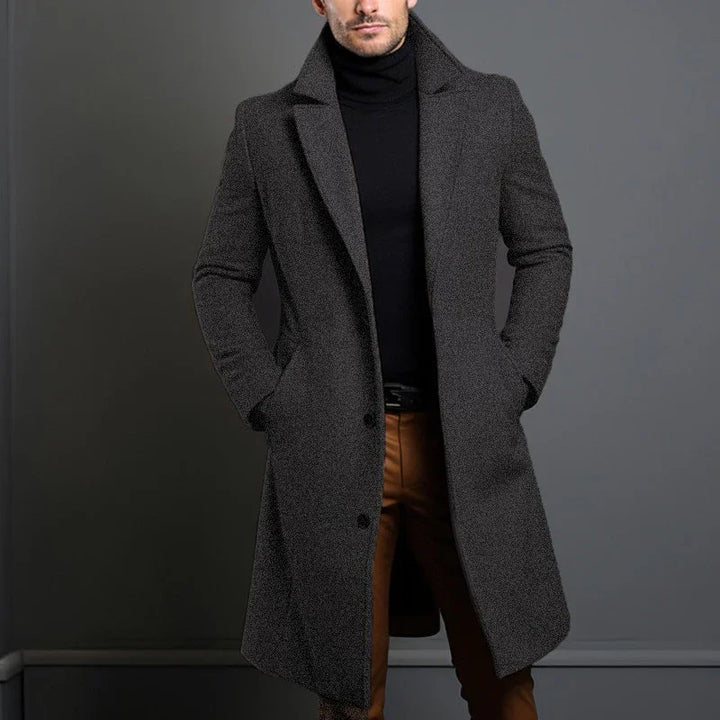 Man wearing dark gray wool coat over black turtleneck and brown pants against gray background