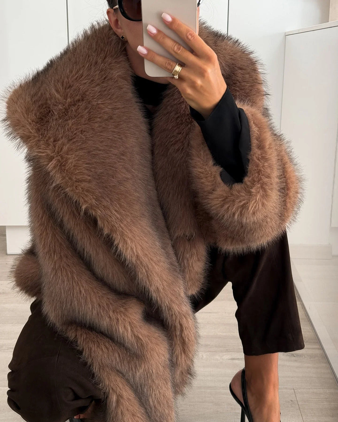 Woman wearing oversized brown fur coat with wide collar and cuffs, black pants, and sunglasses indoors