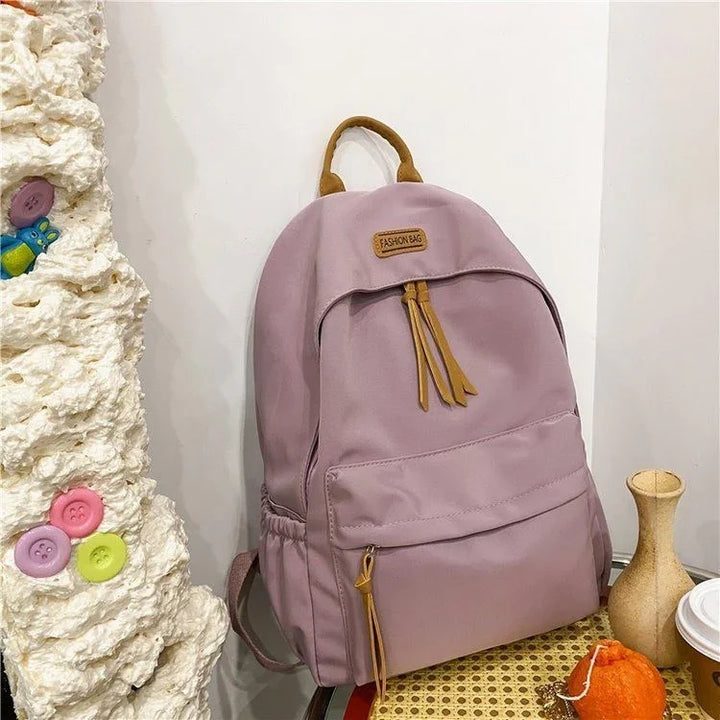 Pink fashion backpack with brown tassels and handle, placed on a chair near textured wall with colorful buttons