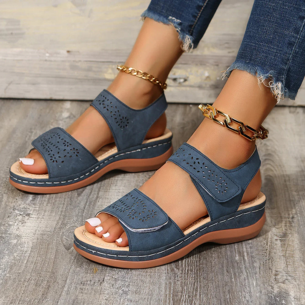 Blue perforated wedge sandals with adjustable straps on feet, paired with gold chain anklets and frayed jeans