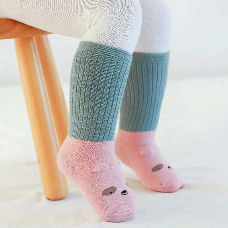 Child wearing ribbed green and pink cotton socks with animal face design while sitting on wooden stool