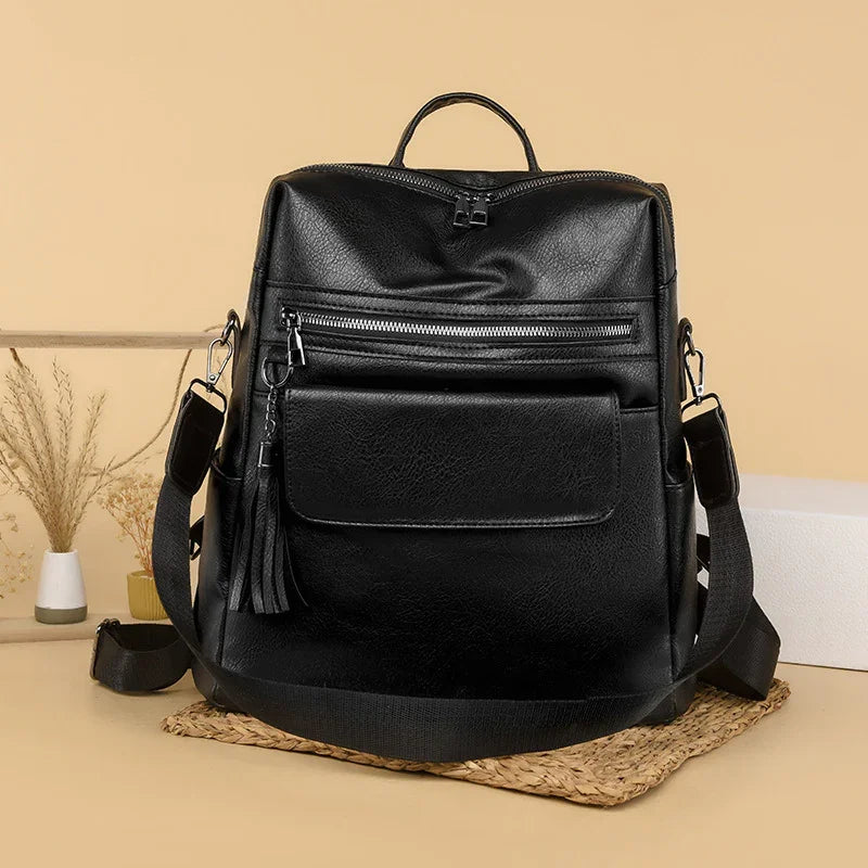 Black leather backpack with front zipper pocket and tassel detail, adjustable straps on beige background
