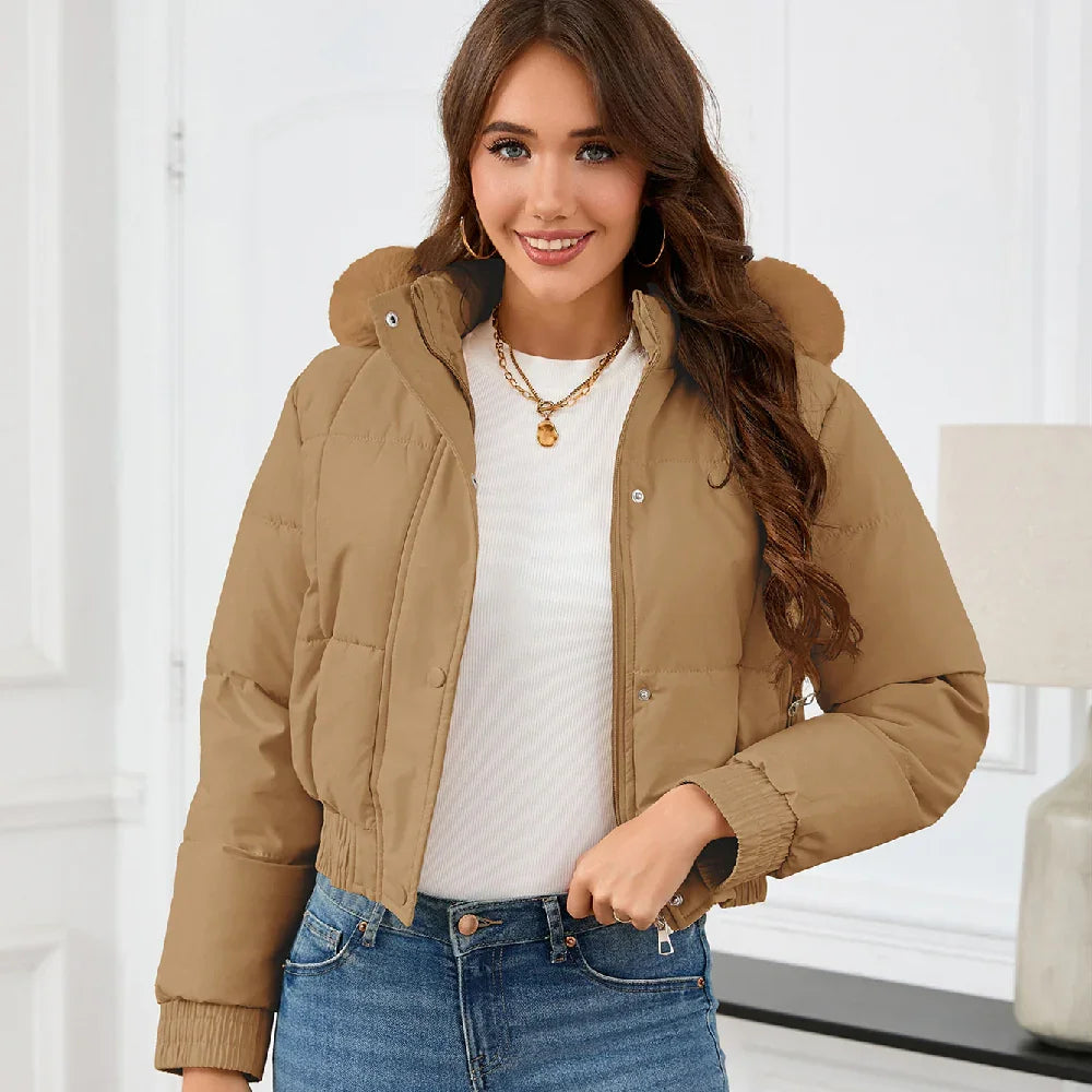 Smiling woman wearing tan cropped puffer jacket with hood, white shirt, and blue jeans indoors
