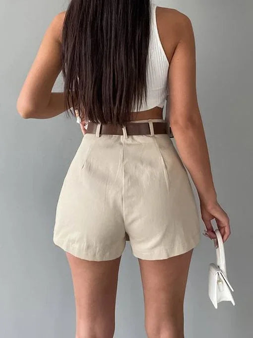 Woman wearing high-waisted beige shorts with brown belt and white crop top holding white handbag