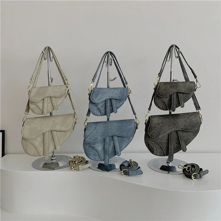 Three sets of saddle-shaped suede handbags in beige, blue, and dark brown displayed on metal stands with matching straps