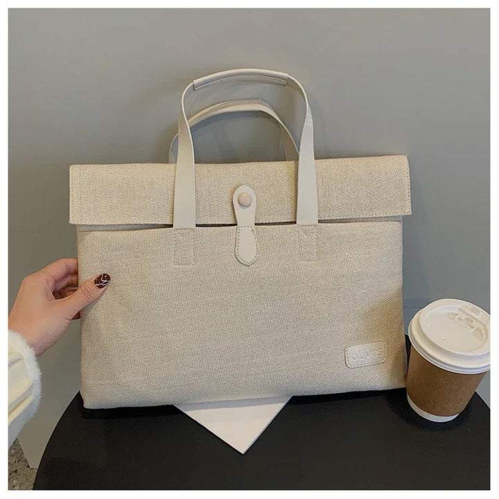 Beige canvas laptop bag with white handles and button closure on black table beside a takeaway coffee cup