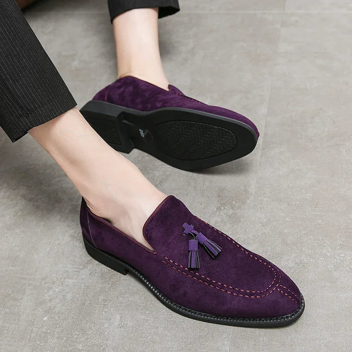 Men's purple suede tassel loafers with black soles worn with black pants on gray floor
