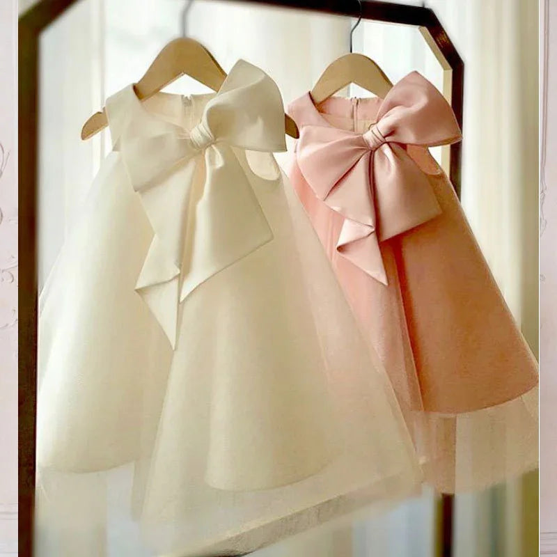 Elegant white and pink girls' party dresses with large bows hanging on wooden hangers