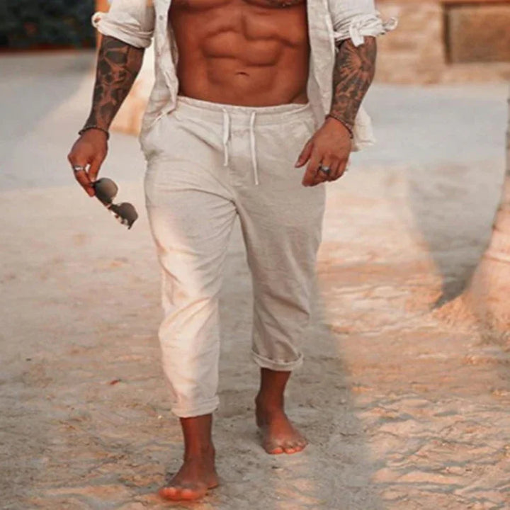 Fit man with tattooed arms wearing rolled-up white pants and open shirt, walking barefoot on sandy ground