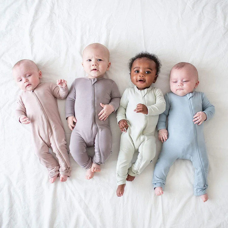 Four diverse babies lying on white blanket wearing pastel zip-up onesies in beige, gray, mint, and blue
