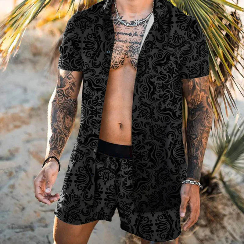 Man wearing black patterned short-sleeve shirt and matching shorts with tattoos on arms and chest on a sandy beach