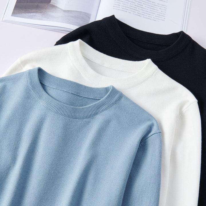 Three folded crewneck sweaters in light blue, white, and black on a white surface with an open book