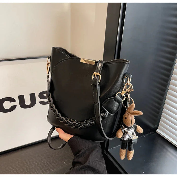 Black leather bucket bag with braided detail and bunny keychain held by hand near window blinds