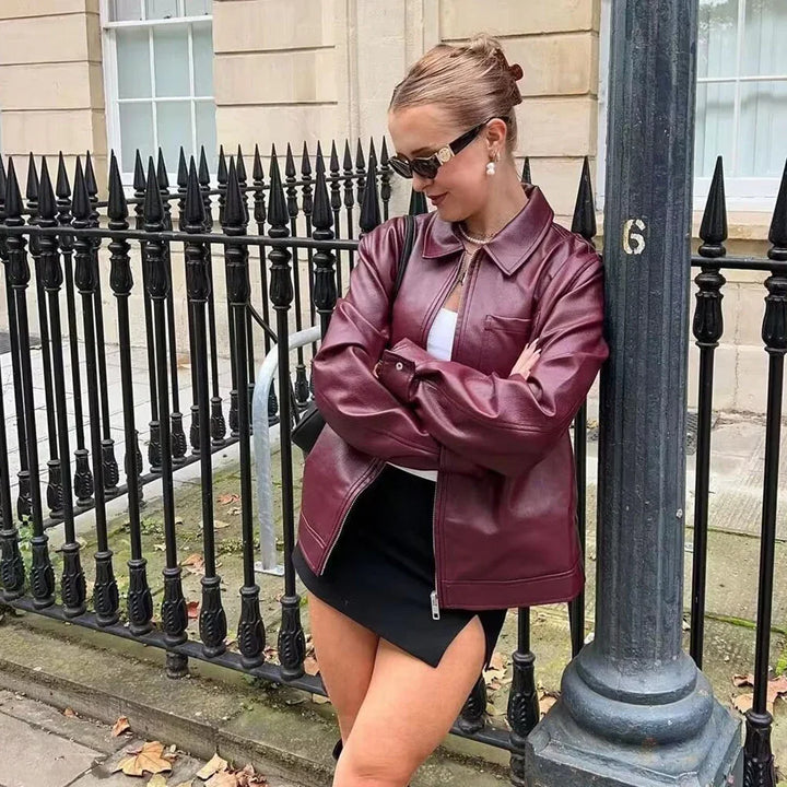 Fashionable woman in maroon leather jacket and black skirt posing by black iron fence on city street