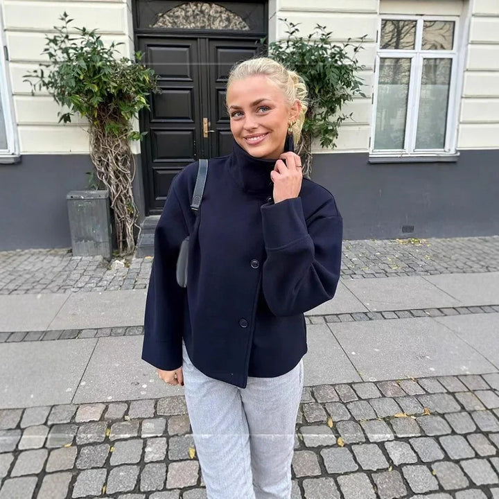 Smiling woman in black jacket and light pants standing on cobblestone street by building with black door