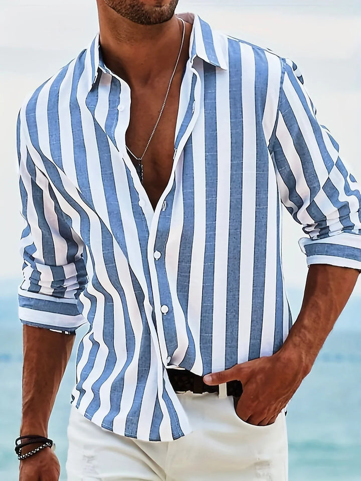 Man wearing blue and white vertical striped casual shirt with rolled-up sleeves and white pants by the ocean