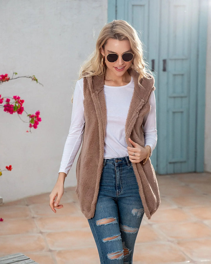 Woman wearing beige fuzzy vest, white long-sleeve shirt, ripped blue jeans, sunglasses, and gold earrings outdoors