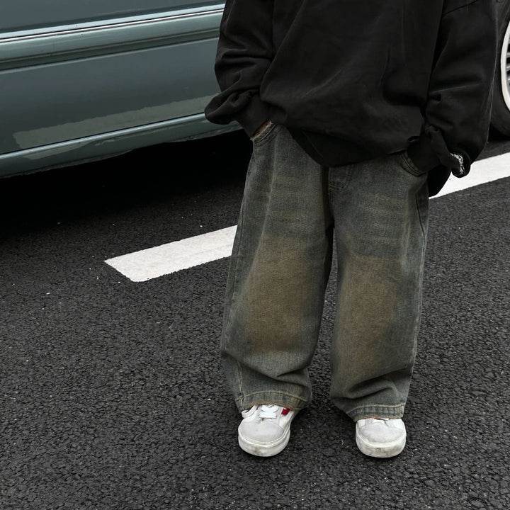 Person wearing oversized baggy jeans, black sweatshirt, and white sneakers standing on asphalt near parked car