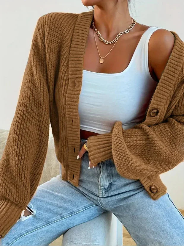 Woman wearing brown chunky knit cardigan, white crop top, light blue ripped jeans, and gold necklaces