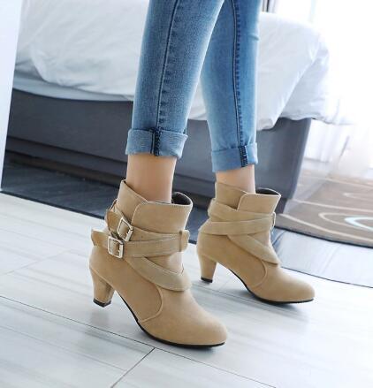 Women's beige ankle boots with decorative buckles and heels worn with rolled-up blue skinny jeans indoors