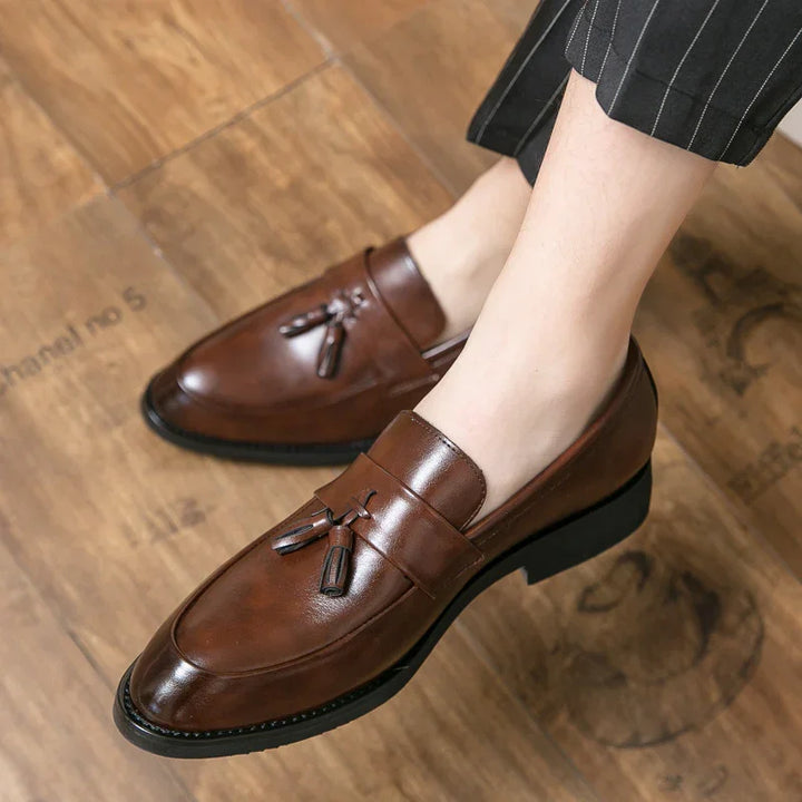 Brown leather tassel loafers worn with black pinstripe pants on wooden floor