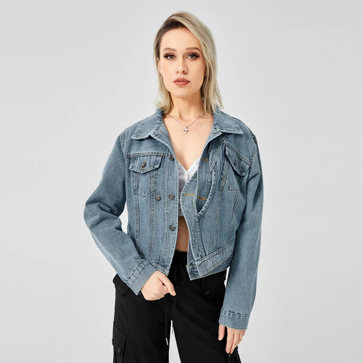 Blonde woman in blue denim jacket, white top, and black pants posing against gray background