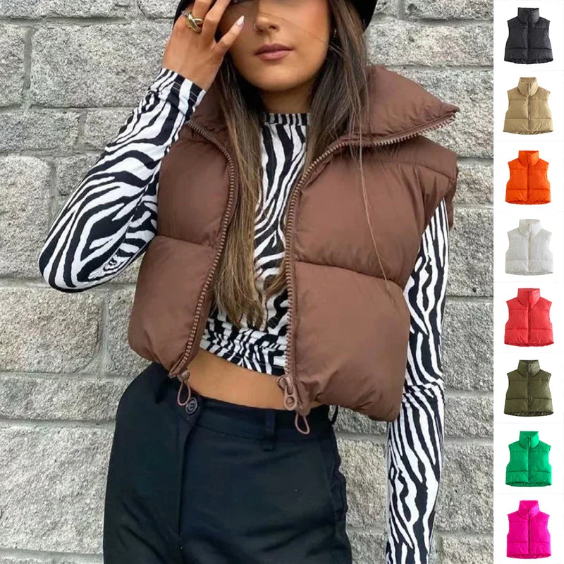 Woman wearing brown puffer vest over zebra print long sleeve top, black pants, and black hat against stone wall